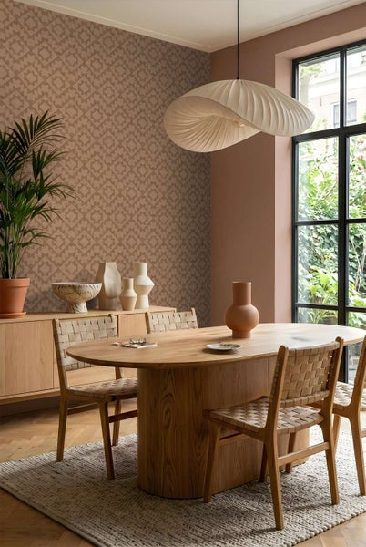 Esszimmer mit gemusterter Tapete, ovalem Holztisch, Stühlen mit Flechtoptik, Sideboard, Dekovasen und einer großen Pendelleuchte am Fenster.