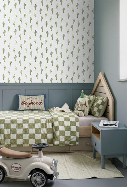 Kinderzimmer mit Kaktus-Tapete, hölzernem Hausbett, karierter Bettwäsche, blauem Nachttisch und Spielzeugauto auf hellem Teppich.