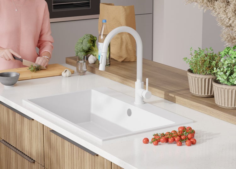 Küchenszene mit weißem Spülbecken und Armatur, Kräutern, Gemüse und Person bei der Essenszubereitung.