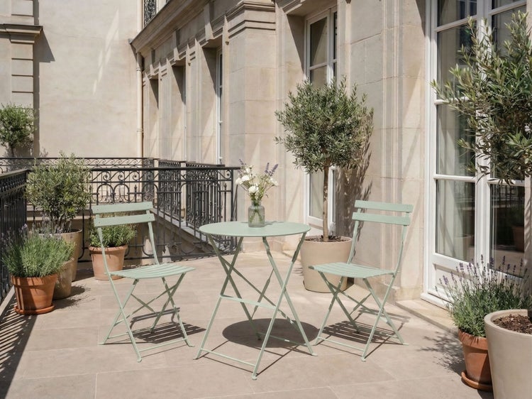 Bistro-Set aus Metall mit einem runden Klapptisch und zwei Klappstühlen auf einer hellen Steinterrasse mit Pflanzen.