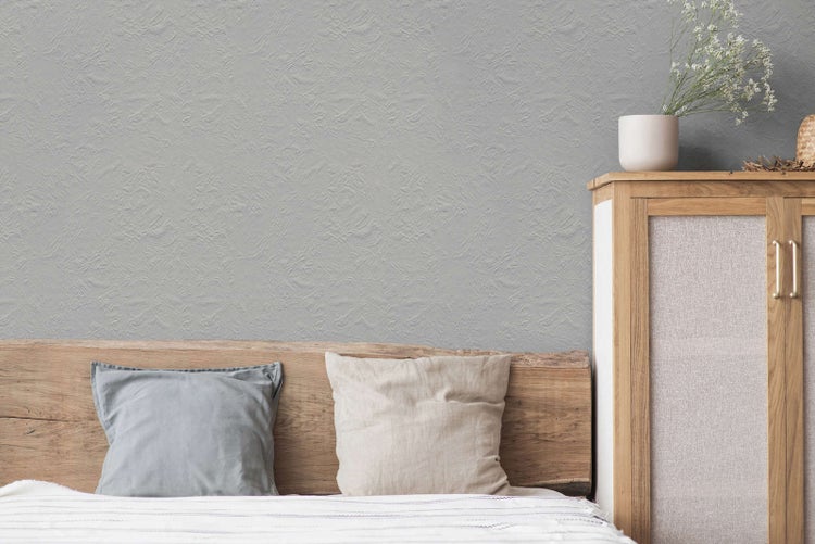 Schlafzimmer mit hellgrauer Strukturtapete hinter einem hölzernen Kopfteil mit Kissen und einem Holzschrank.