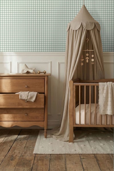 Kinderzimmer mit Holzkommode, Gitterbett mit beigem Betthimmel und grün-weiß karierter Tapete über weißer Wandtäfelung.