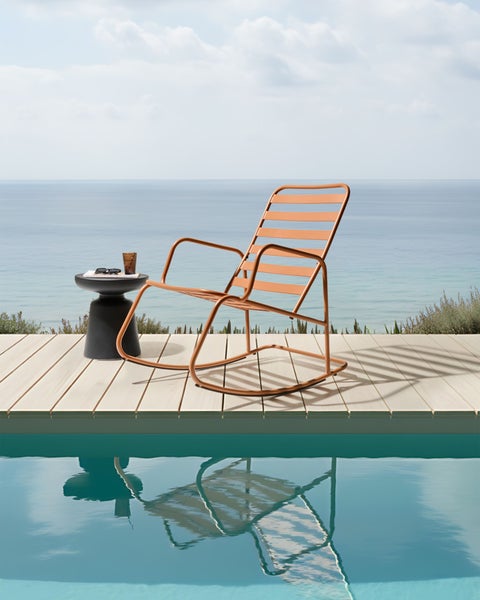 Szene mit einem orangefarbenen Schaukelstuhl aus Metall, einem schwarzen Beistelltisch und Blick auf das Meer