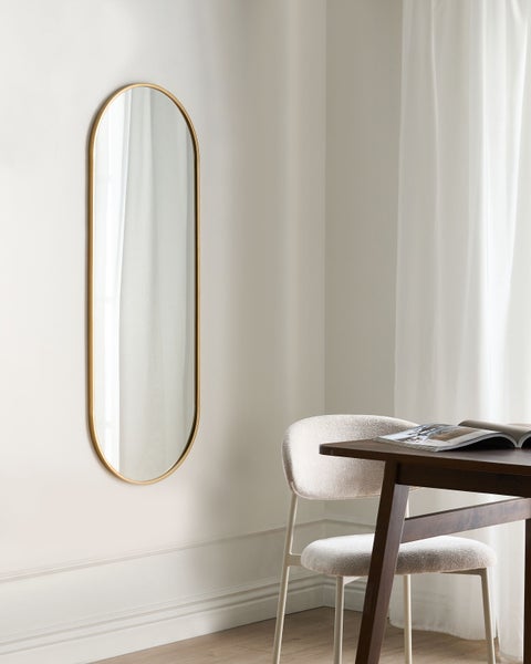 Ein langer, ovaler Wandspiegel mit schmalem goldfarbenem Rahmen in einem hellen, modern eingerichteten Raum neben einem Tisch.