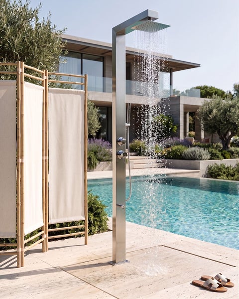Moderne Außendusche aus Edelstahl mit Regenduschkopf und Handbrause auf einer Steinterrasse neben einem Pool und einem Bambus-Sichtschutz.