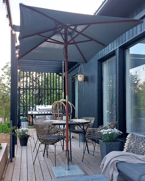 Helle, moderne Terrasse mit großem Sonnenschirm, Esstisch, Rattansesseln und Hängesessel auf einem Holzdeck neben einem Holzhaus mit Gasgrill.