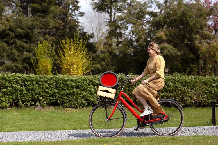 Eine Frau fährt auf einem roten Fahrrad mit einer Holzkiste am Frontgepäckträger auf einem Kiesweg in einem hellen Garten.