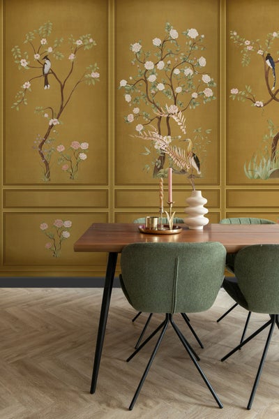 Ein Esszimmer mit einem Holztisch, grünen Stühlen und einer gemusterten Tapete mit Vogel- und Blumenmotiven.