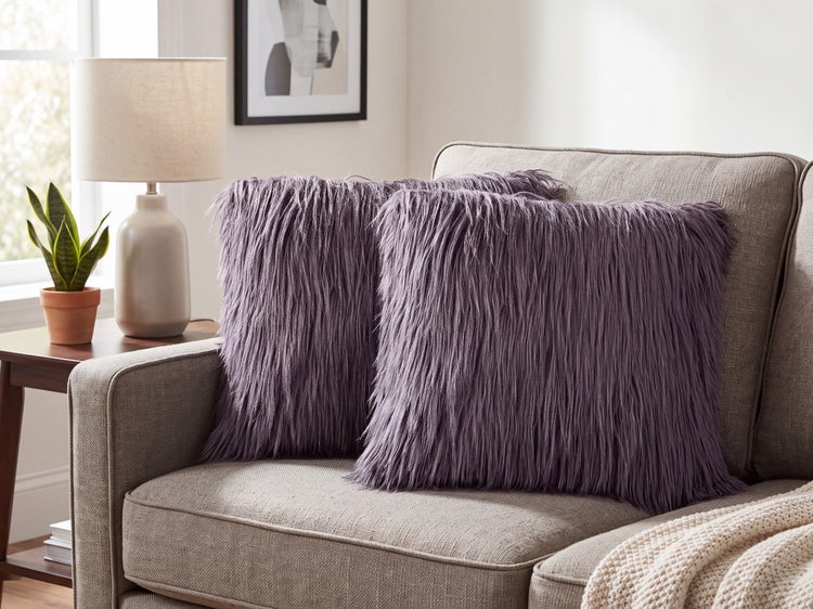 Zwei flauschige Dekokissen mit langem Flor auf einem modernen Sofa mit Beistelltisch, Lampe und Pflanze im Hintergrund.