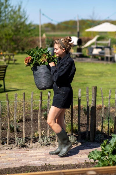 Eine Frau trägt einen großen, strukturierten dunkelgrauen Pflanztopf mit einer blühenden Hortensie in einem Garten an einem Staketenzaun.