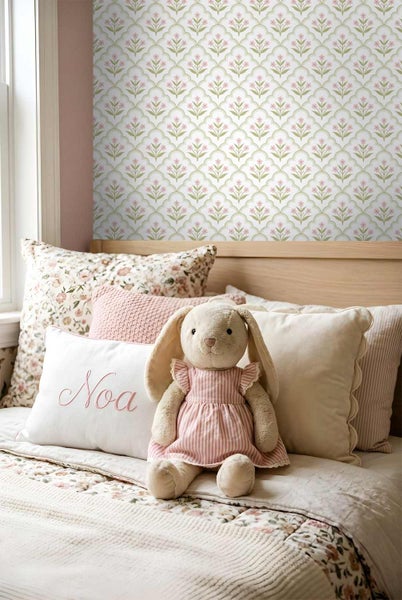 Kinderzimmer mit Tapete mit rosa Blumenmuster in geschwungenen grünen Rahmen, Holzbetthaupt, Kissen und einem Stoffhasen.