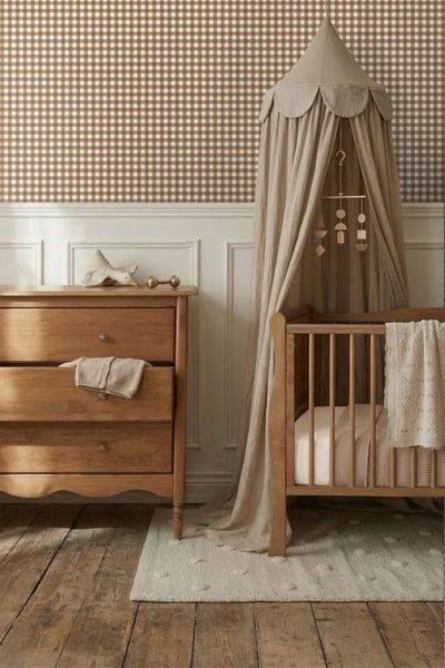 Kinderzimmer mit braun-weiß karierter Vliestapete, weißer Wandvertäfelung, Holzkommode und einem Babybett aus Holz mit Stoffhimmel.