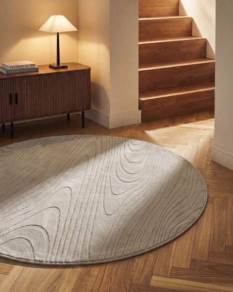 Runder Teppich mit Wellenmuster auf Fischgrätparkett neben einer Holztreppe und einem Sideboard mit Lampe.