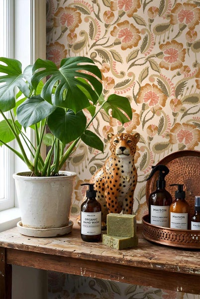 Vliestapete mit großem floralem Muster hinter einer Monstera-Pflanze, einer Leopardenfigur und Badaccessoires auf einem rustikalen Holztisch.