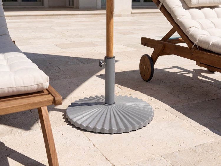 Geriffelter Sonnenschirmständer auf einer hellen Steinterrasse zwischen zwei hölzernen Gartenliegen mit hellen Polstern in einer hellen Umgebung.