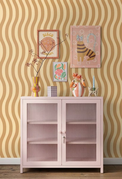 Vliestapete mit vertikalem Wellenmuster in Ocker und Beige in einem modern eingerichteten Zimmer mit rosa Metallschrank und Dekoration.