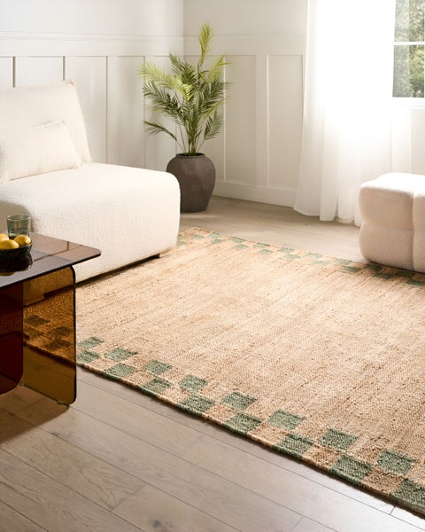 Gewebter Teppich aus Jute mit grünem Karomuster am Rand in einem hellen Wohnzimmer mit weißen Polstermöbeln und Holzfußboden.