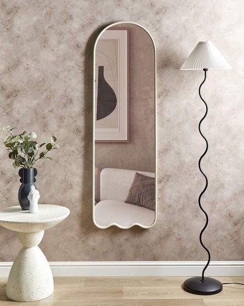 Zimmer mit oben gewölbtem Wandspiegel mit Wellenkante unten, schwarzer Stehlampe mit geschwungenem Fuß und weißem skulpturalem Beistelltisch.