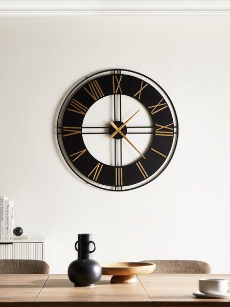 Große runde Wanduhr aus Metall mit goldfarbenen römischen Ziffern über einem Holztisch in einem hellen, modernen und organisierten Raum.