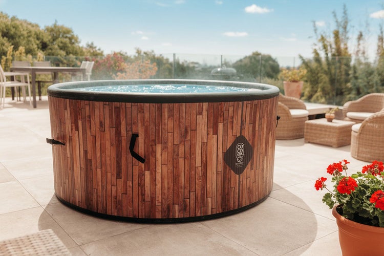 Runder Whirlpool mit Verkleidung in Holzoptik und Coco Spa Logo auf einer hellen Terrasse mit Gartenmöbeln.