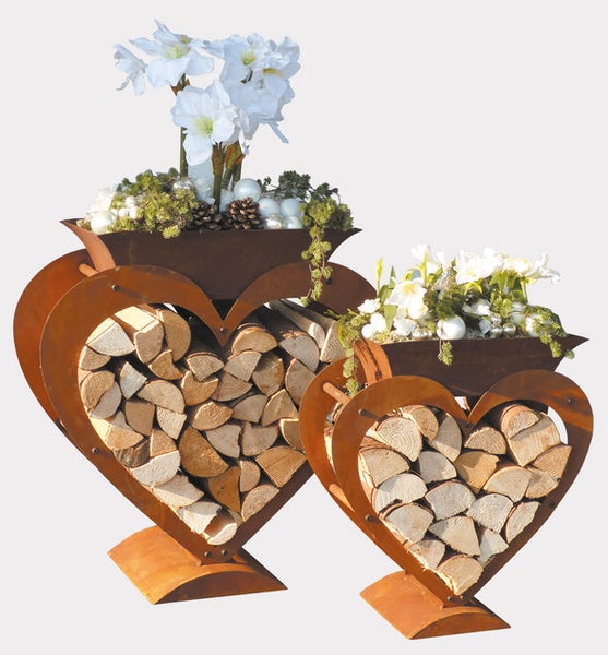 Herzförmiges Kaminholzregal-Set aus Metall in Rostoptik, befüllt mit Brennholz und einer Pflanzschale mit weißen Blumen.