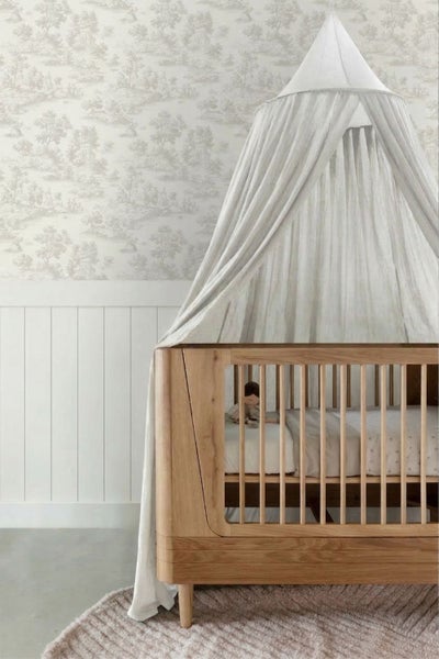 Kinderzimmer mit Babybett aus Holz, Stoffbaldachin und Tapete mit Landschaftsmuster über weißer Wandvertäfelung.