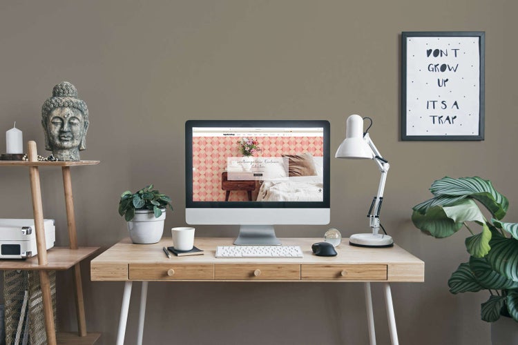 Moderner Arbeitsplatz mit hellem Holzschreibtisch, Computer, weißer Schreibtischleuchte und Zimmerpflanzen vor taupefarbener Wand.