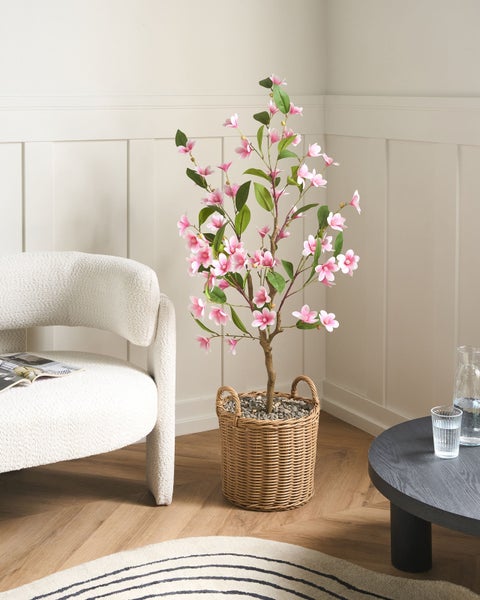 Künstliche Magnolie mit rosa Blüten in einem geflochtenen Korb, neben einem weißen Sessel in einem hellen Wohnzimmer mit Holzfußboden.