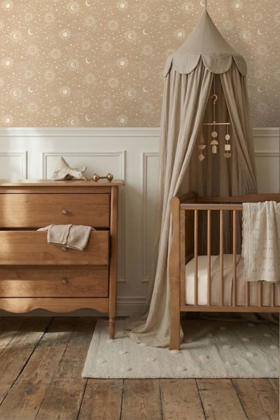 Kinderzimmer mit Tapete mit Sonnen- und Mondmuster, Holzgitterbett mit Baldachin und Holzkommode vor Wandvertäfelung und Holzfußboden.