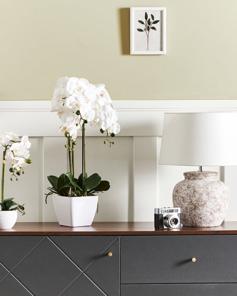 Dekoratives Arrangement mit künstlichen Orchideen, Tischlampe und Vintage-Kamera auf einer Kommode.