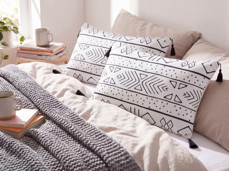 Bett mit zwei rechteckigen Dekokissen mit schwarzem geometrischem Muster und Quasten auf beiger Bettwäsche mit grauer Strickdecke.