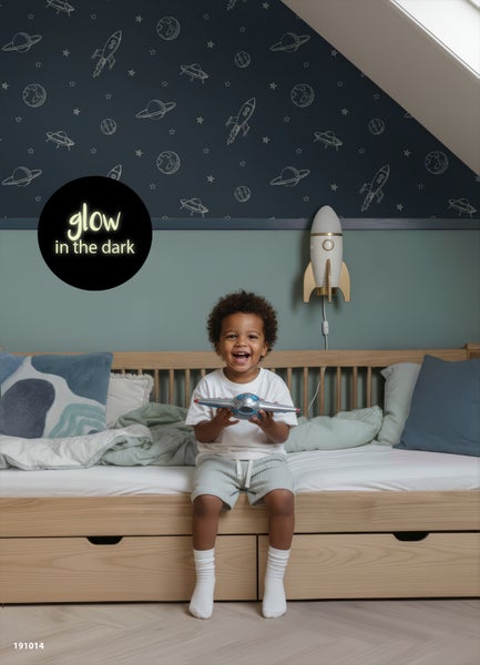 Helles Kinderzimmer mit im Dunkeln leuchtender Weltraumtapete und Holzbett. Ein Kind spielt auf dem Bett. Eine Raketenlampe hängt an der Wand.