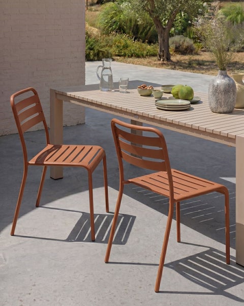 Zwei orangefarbene Gartenstühle aus Metall an einem hellen Gartentisch mit Lattenstruktur auf einer sonnigen Terrasse.