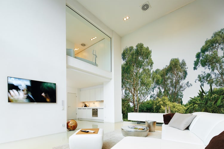 Modernes, helles Wohnzimmer mit großer Fototapete mit Waldmotiv, weißem Sofa, silbernen Tischen und offener Küche im Hintergrund.