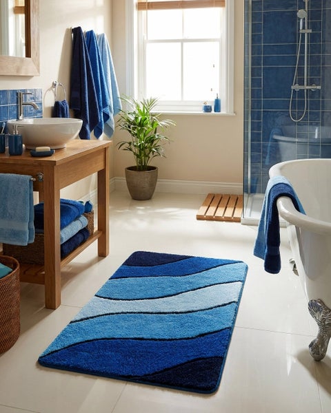 Helles Badezimmer mit blauem Badeteppich im Wellendesign, Waschtisch aus Holz, weißen Sanitärobjekten und verschiedenen blauen Handtüchern.