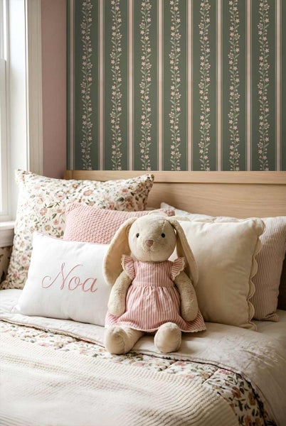 Kinderzimmer mit Vliestapete mit vertikalen Streifen und Blumenranken hinter einem hellen Holzbett mit verschiedenen Kissen und einem Stoffhasen.