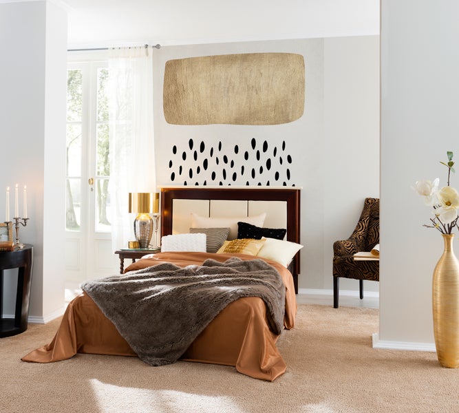 Helles Schlafzimmer mit Bett, brauner Tagesdecke, Pelzpläd, gemustertem Sessel, goldener Bodenvase und Wandgestaltung in Gold und Schwarz.