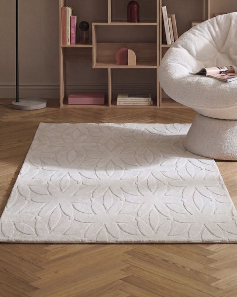 Ein rechteckiger Teppich mit Blumenmuster auf einem Parkettboden in einem modern eingerichteten Raum mit Bücherregal und Loungesessel.