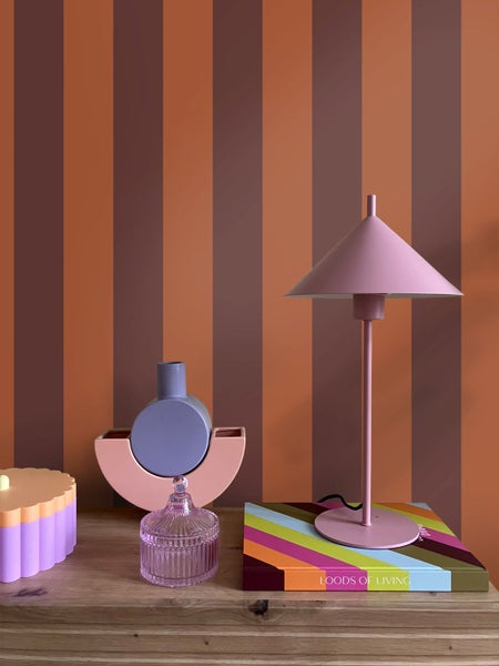 Modernes Interieur mit gestreifter Tapete, einer Tischleuchte mit kegelförmigem Schirm, geometrischen Vasen und einer Glasdose auf einer Holzfläche.