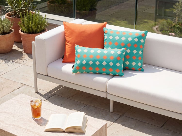 Modernes Lounge Sofa mit Dekokissen in geometrischem Design auf einer sonnigen Terrasse mit Pflanzkübeln und einem Beistelltisch.
