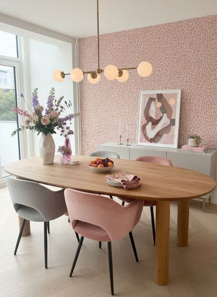 Helles Esszimmer mit ovalem Holztisch, rosa Tapete mit Leopardenmuster, Polsterstühlen und einer modernen mehrflammigen Kugelleuchte.