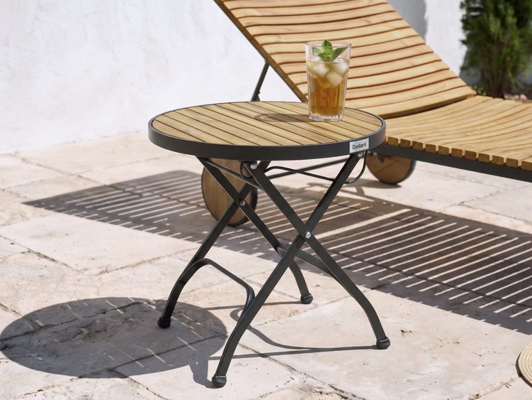 Runder Gartenbeistelltisch mit Holzlattenplatte und schwarzem Klappgestell aus Metall auf einer hellen Terrasse neben einer Liege. Beliani Logo.