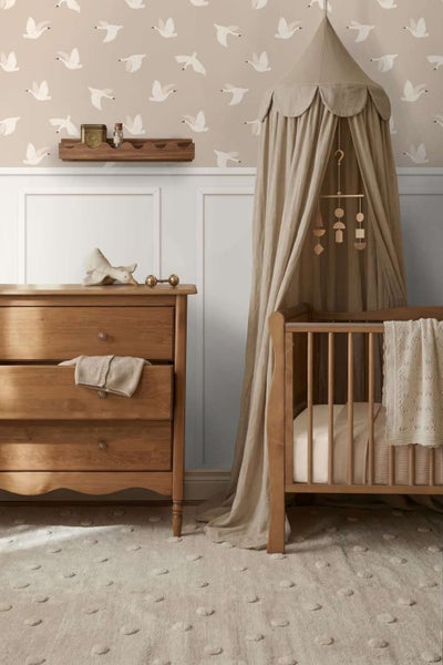 Ein gemütliches Babyzimmer mit Holzmöbeln, einer Kommode, einem Gitterbett mit Betthimmel und einer Vliestapete mit Schwanenmuster.