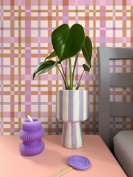 Vliestapete mit buntem Karomuster hinter einer Monstera Pflanze in einer gestreiften Vase und einer lila Kerze.