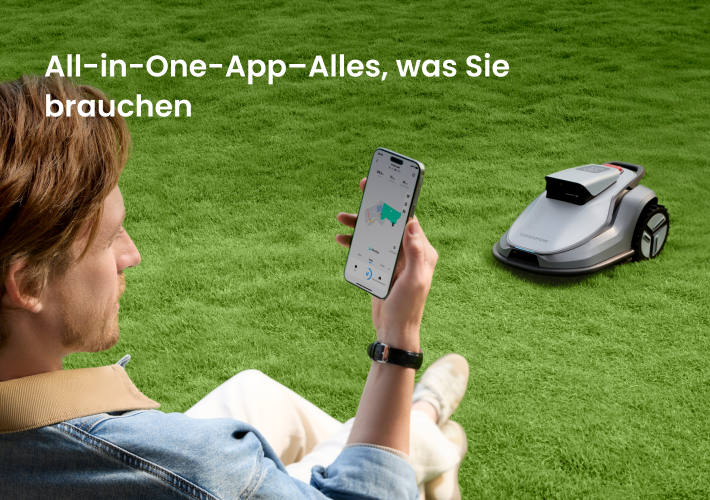 Ein Mann sitzt auf einem Rasen und steuert einen silbernen Mähroboter mit einer App auf seinem Smartphone.