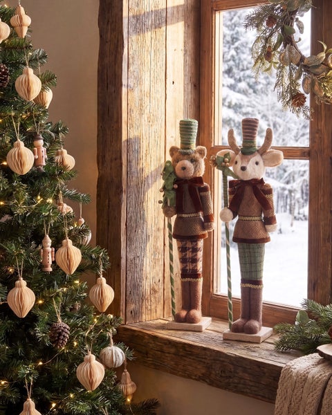 Festliche Weihnachtsdekoration mit zwei stehenden Stofffiguren, Bär und Rentier, auf einer rustikalen Holzfensterbank neben einem Tannenbaum.