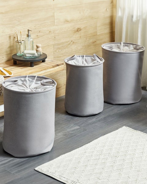 Drei graue Wäschekörbe aus Stoff mit Kordelzug in einem hellen, organisierten Badezimmer mit Holzwand und grauem Boden.
