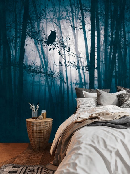 Fototapete mit einem mystischen blauen Wald und einer Eule auf einem Ast, in einem Schlafzimmer mit Bett und Beistelltisch aus Geflecht.