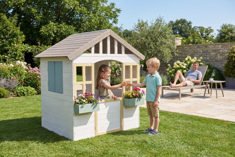 Kinderspielhaus aus Holz in Weiß und Grau mit Blumenkästen in einem sonnigen Garten mit spielenden Kindern und einer Sonnenliege.