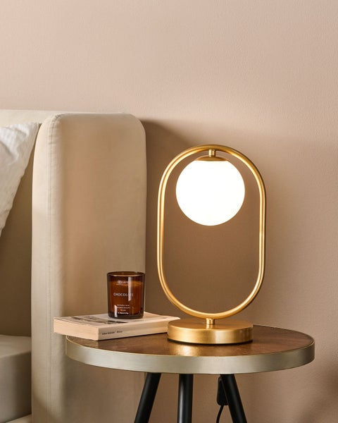 Moderne goldfarbene Tischleuchte mit weißem kugelförmigem Schirm auf einem runden Beistelltisch neben einem gepolsterten Bett.